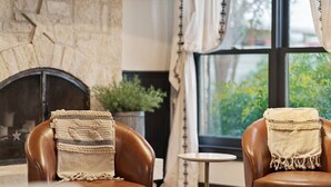 A room with two leather armchairs, a small table, a stone fireplace, and a view through a large window. - Stonewall Main Haus - Sleeps 6 (Stonewall)