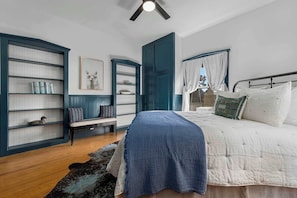 Un dormitorio con cama, estantería, banca, ventana con cortinas y ventilador de techo.