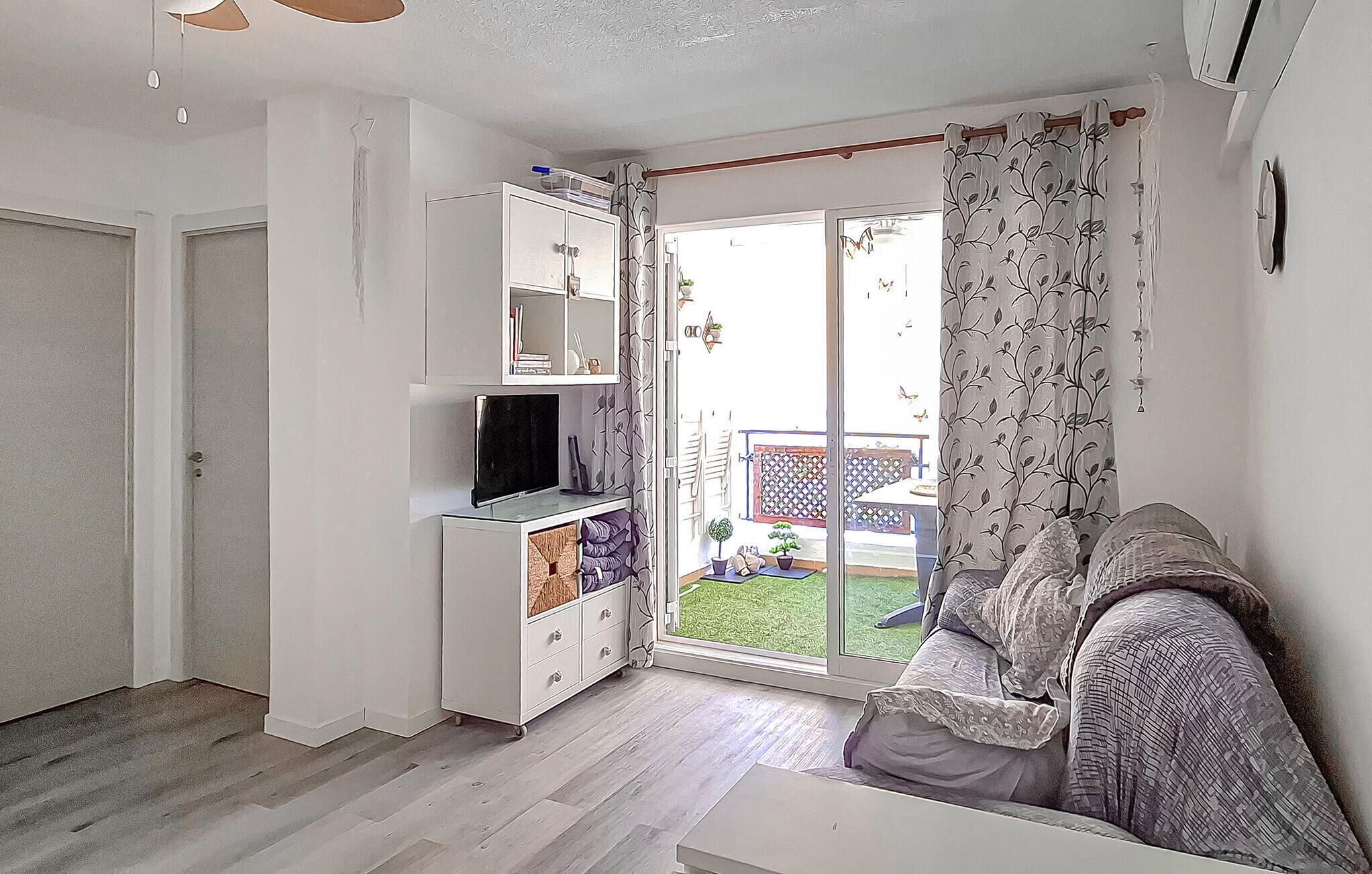 Una sala de estar con sofá, televisor sobre un mueble bajo, puerta corrediza de vidrio a un balcón y un armario blanco con estantes.