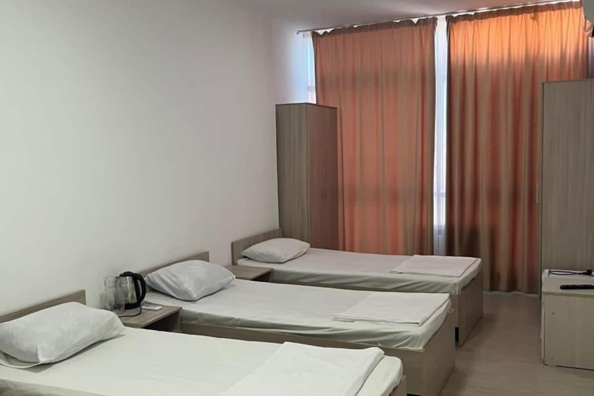 A hotel room with three single beds, a wooden wardrobe, a small desk, and a window with curtains.