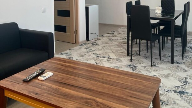 A modern living room with a wooden coffee table, a black sofa, a dining area with a table and chairs, and a television mounted on the wall. - Zeraviktoria (Atyrau)