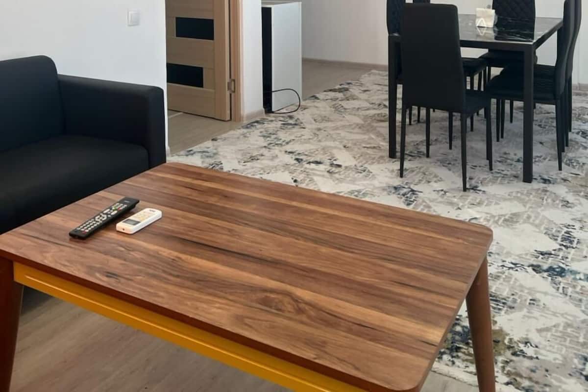 A modern living room with a wooden coffee table, a black sofa, a dining area with a table and chairs, and a television mounted on the wall.