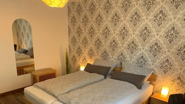 A double bed with patterned bedding, a nightstand with a lamp, a mirror, and a patterned wallpaper.