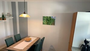 A dining area with a wooden table, green upholstered chairs, a hanging light fixture, a framed picture on the wall, and a coffee maker in the background.