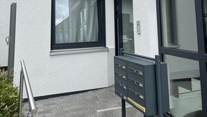 Fachada de edificio con varias ventanas, una reja metálica y un pequeño espacio exterior con un mueble metálico.