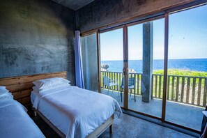 Un dormitorio con dos camas, un balcón con vista al mar y una pared de concreto.