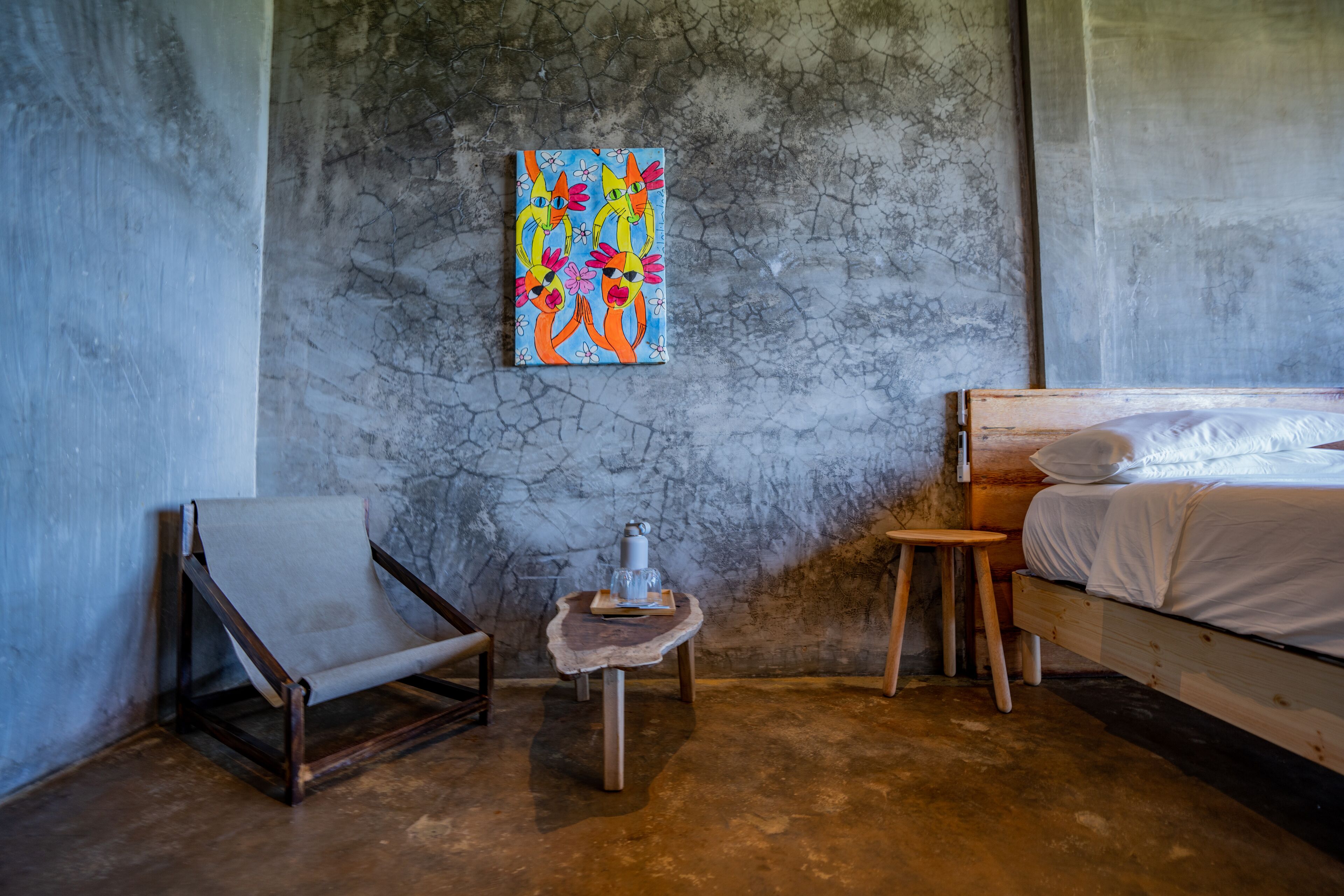 Een kamer met een bed, een stoel, een klein tafeltje met een fles en glazen, en een kleurrijk schilderij aan de muur.