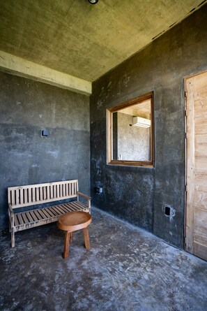 Una habitación con piso de concreto, un banco de madera, un taburete pequeño y una ventana con marco de madera.
