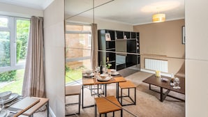 A modern dining area with a table set for four, a bookshelf, and a view of the garden through a large window. - Stunning 2-bed Apartment in Windsor - Free Parking (Windsor)