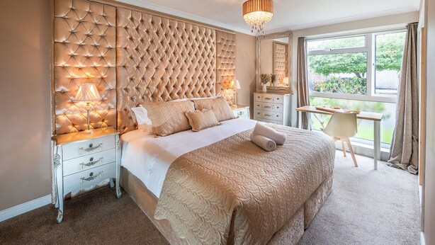 A bedroom with a tufted headboard, a large bed with a beige bedspread, a nightstand with a lamp, and a desk with a chair. - Stunning 2-bed Apartment in Windsor - Free Parking (Windsor)
