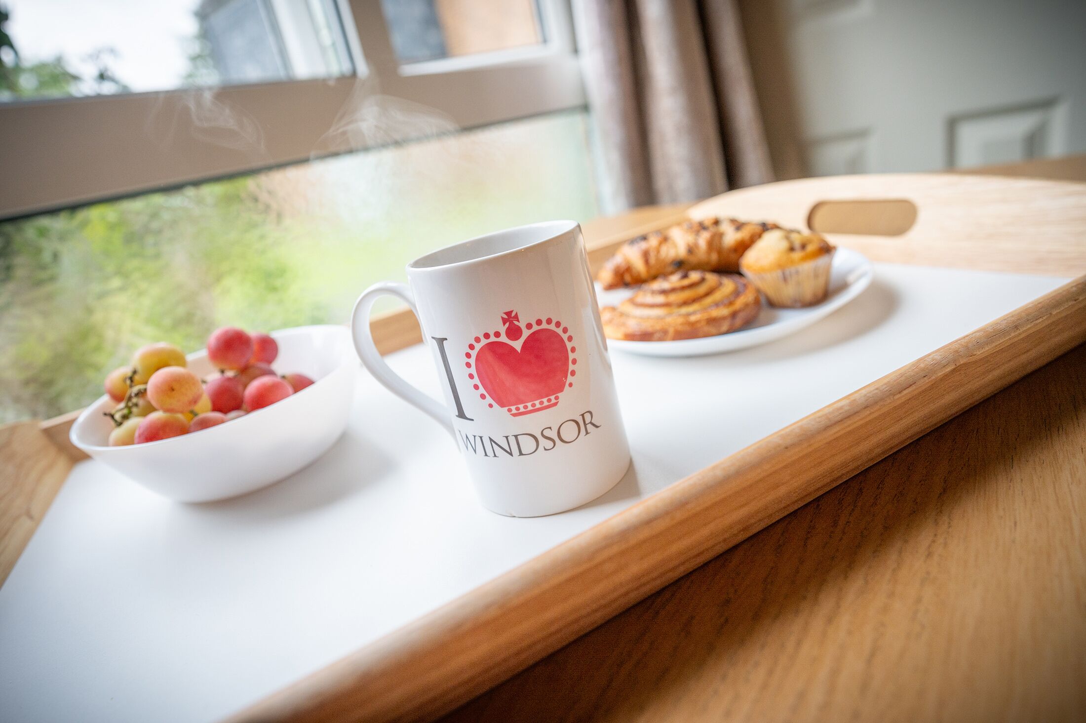 Una bandeja con una taza, un bol de fruta y pasteles sobre una mesa cerca de una ventana.