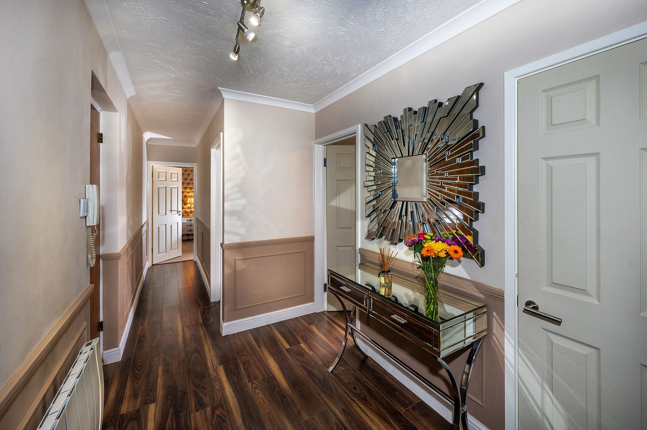A hallway with wooden flooring, a mirror with a sunburst design, and a vase with flowers on a console table.