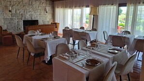 A restaurant with stone walls, wooden ceiling, and large windows with curtains. Tables are set with white tablecloths and silverware.