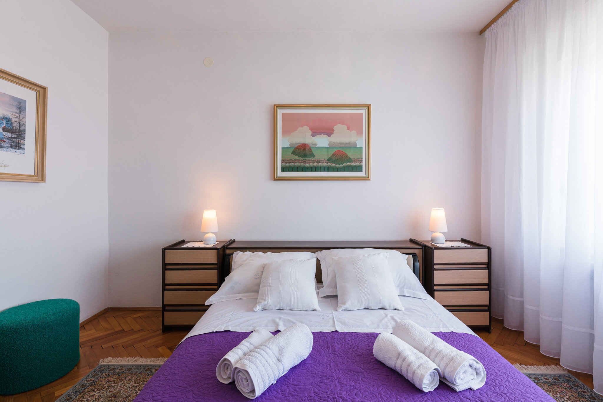 Una cama bien hecha con una colcha morada y almohadas blancas, dos lámparas de noche y un cuadro enmarcado en la pared.