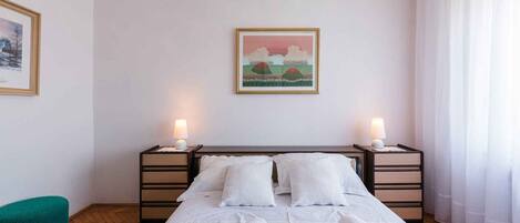 Una cama bien hecha con una colcha morada y almohadas blancas, dos lámparas de noche y un cuadro enmarcado en la pared.