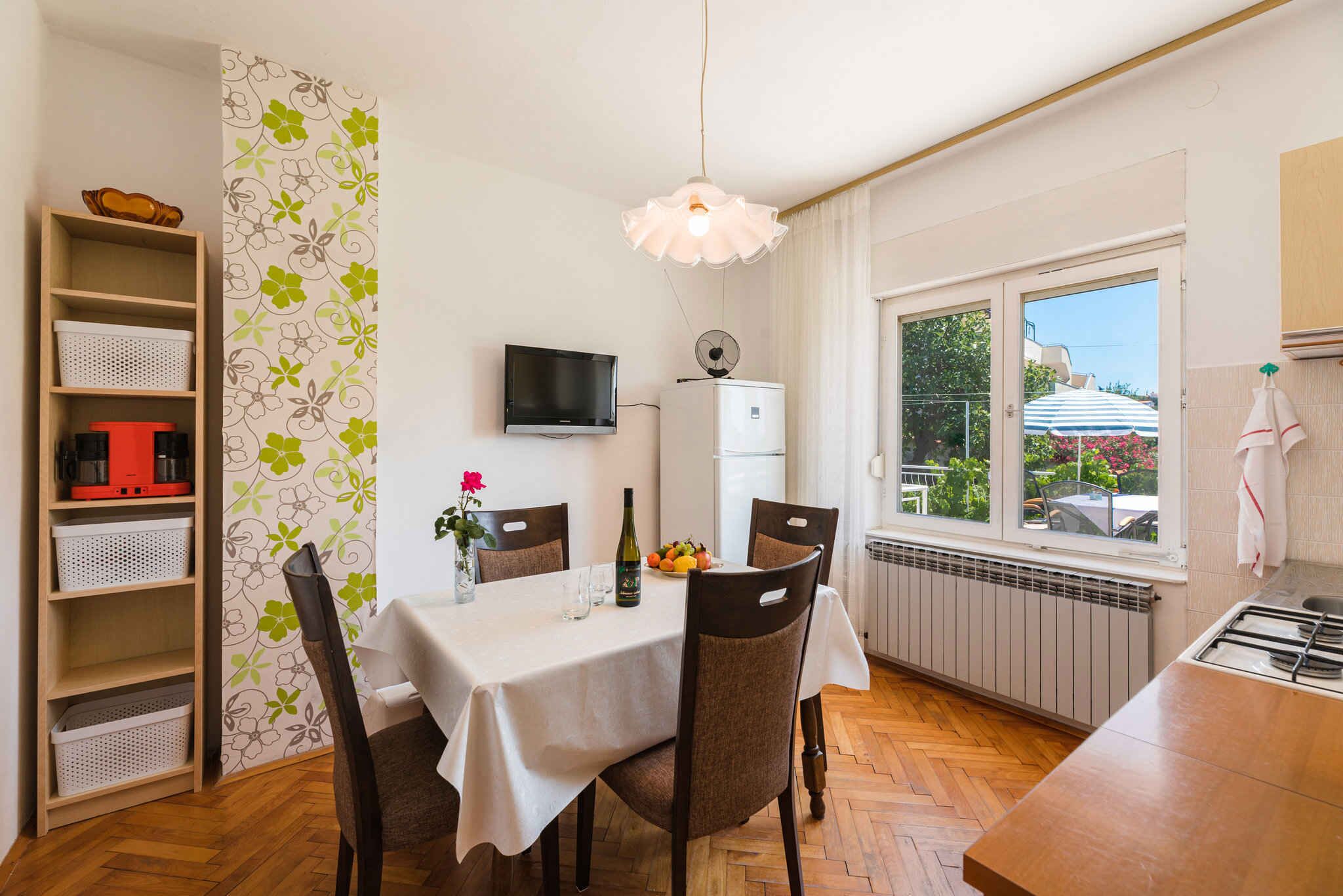 A dining area with a table set for two, a television mounted on the wall, a radiator, and a window with a view of the outdoors.