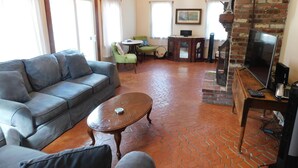 A living room with a grey sofa, a wooden coffee table, and a TV on a wooden stand. There is a brick wall and a window with curtains. - Path to Pond, Private Location, Sweet Home! (CS0031) (Wellfleet)