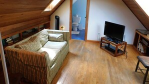 A cozy attic room with a wooden ceiling, a striped sofa, a television on a wooden stand, and a view of the bathroom through an open door. - Path to Pond, Private Location, Sweet Home! (CS0031) (Wellfleet)