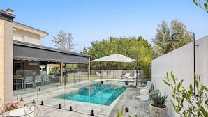 Un espace piscine extérieur moderne, doté d’un coin salon couvert, de chaises longues et de plantes en pot.
