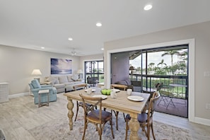 A spacious living room with a large dining table, comfortable seating, and a view of the outdoors through large windows.