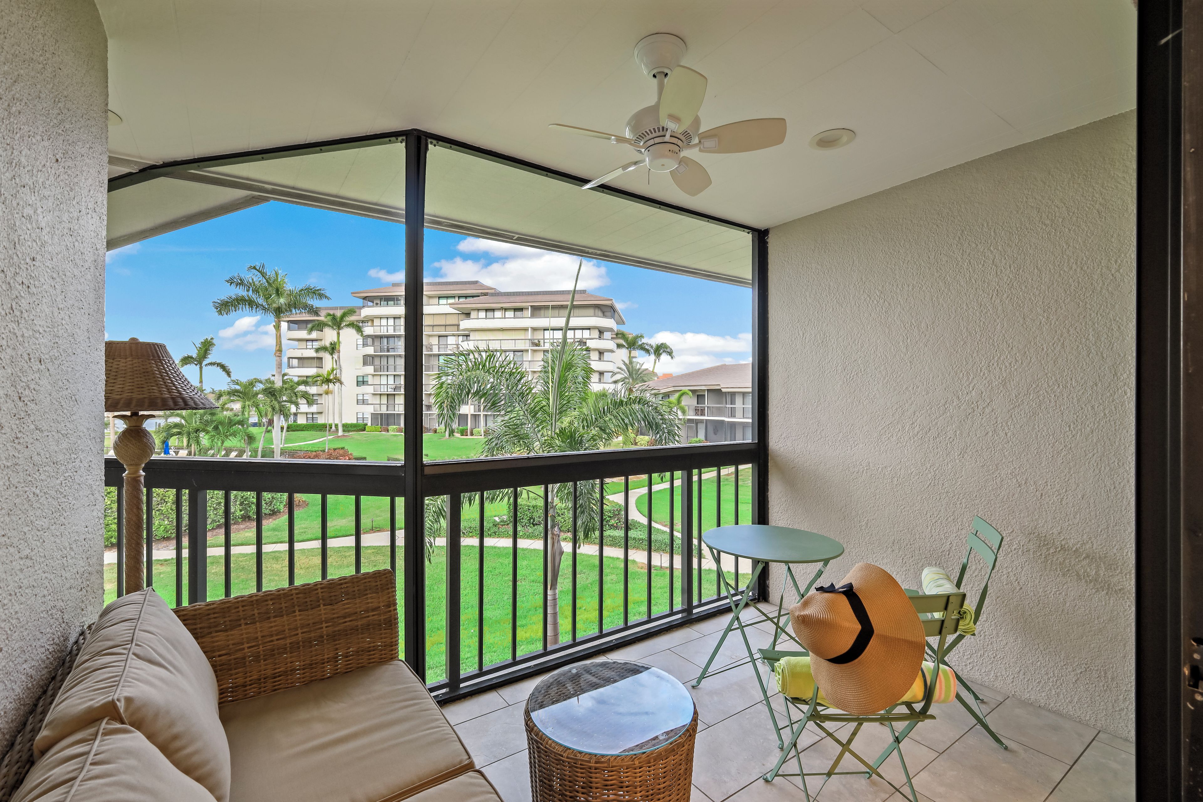 A balcony with a sofa, a small table, and a chair. There is a view of a building and palm trees in the background.