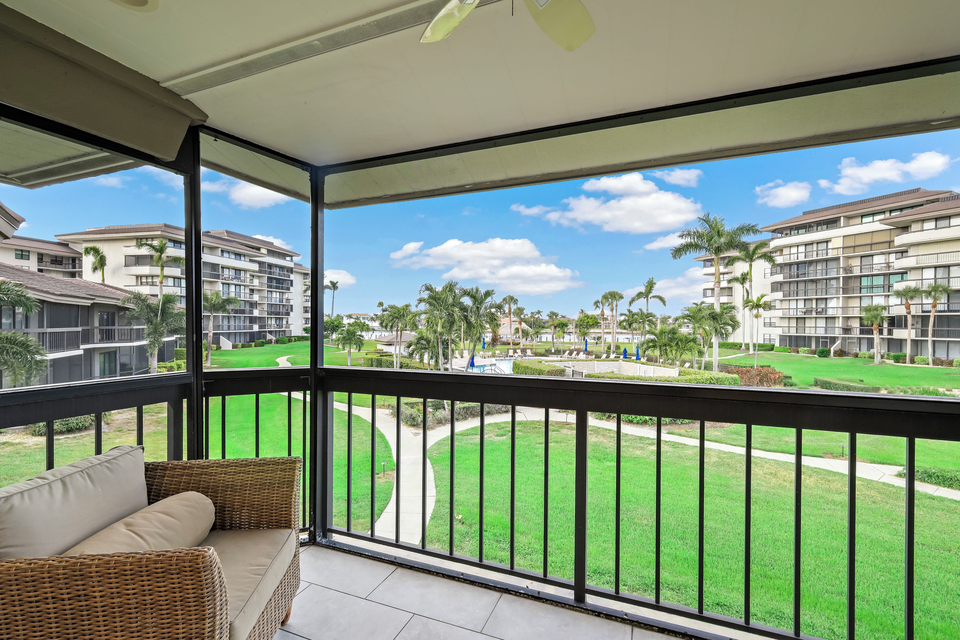 A balcony with a wicker sofa and a view of a well-maintained lawn and buildings.