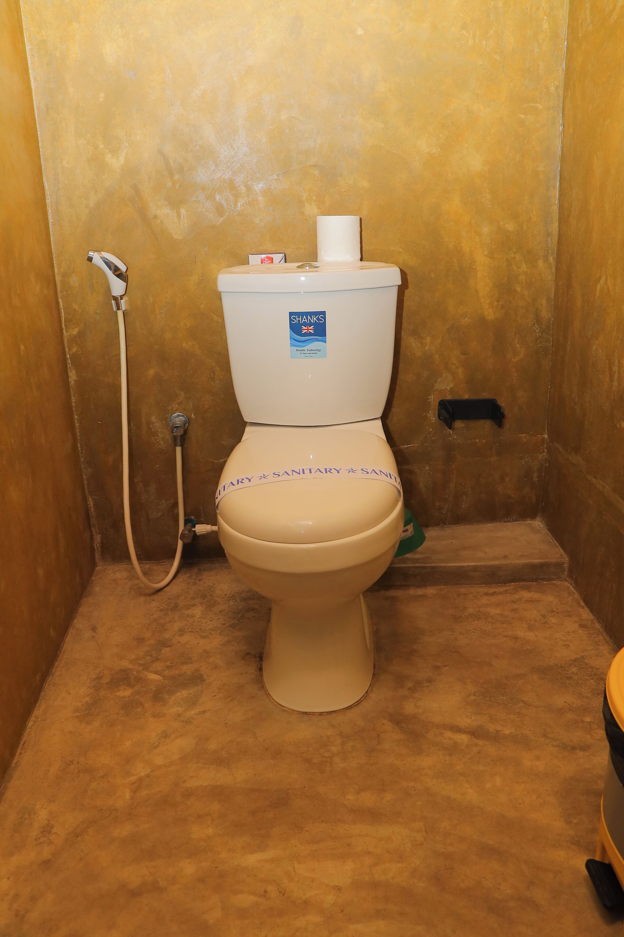 A toilet with a "SHAMKS" label, a toilet paper holder, and a shower head.