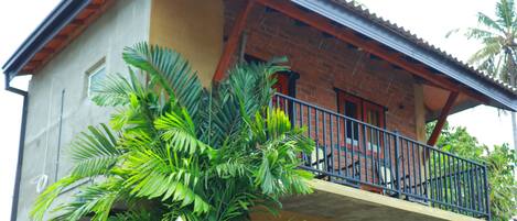 Un edificio de dos pisos con balcón, pared de piedra y plantas tropicales.
