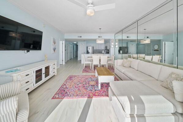 A modern living room with a white sofa, a coffee table, and a TV on the wall. There is a kitchen in the background with white cabinets and a dining area with white chairs.