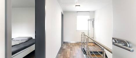 Une chambre d’hôtel moderne avec un lit superposé, un plancher en bois, un escalier doté d’une rampe en métal et une grande fenêtre laissant entrer la lumière naturelle.