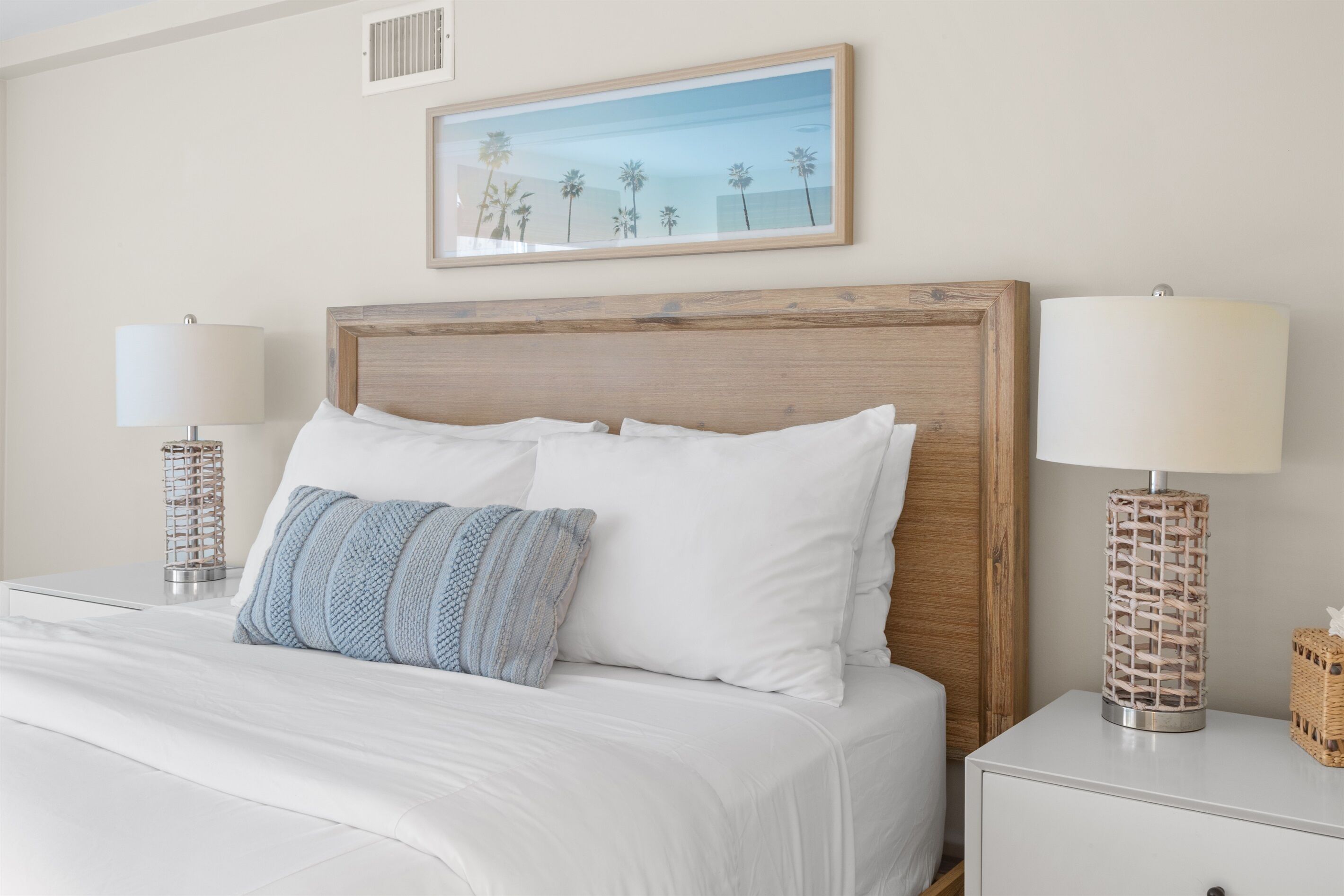 A neatly made bed with white linens and a blue and white pillow, flanked by two bedside tables with lamps and a framed picture above.
