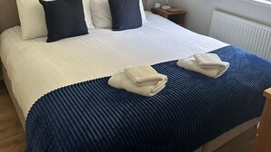 Una cama bien hecha con una colcha azul y blanca, dos almohadas y dos toallas dobladas sobre la cama. Encima de la cama, en la pared, hay tres cuadros enmarcados.
