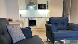 Una sala moderna con un sofá gris oscuro, un sofá azul y una mesa de centro de vidrio. Se ve una cocina con gabinetes blancos y una campana extractora de acero inoxidable al fondo.