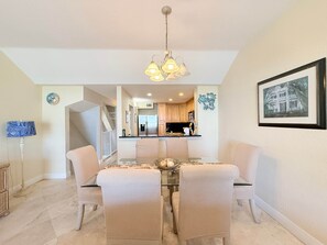 A dining area with a glass table, cushioned chairs, a chandelier, and a kitchen in the background. - Oceanview • Pool View • Indoor/Outdoor Pickleball (Key Largo)