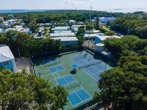 An aerial view of a residential area with multiple tennis courts, houses, and a large body of water in the background. - Oceanview • Pool View • Indoor/Outdoor Pickleball (Key Largo)