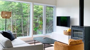 A modern living room with a sofa, armchair, and a TV. A fireplace is visible, and the room has large windows offering a view of trees. - Lakefront cottage in the Laurentians (Saint-Adolphe-d'Howard)