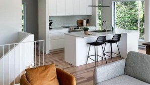 A modern kitchen with white cabinetry, a central island with bar stools, and a large window providing a view of greenery outside. - Lakefront cottage in the Laurentians (Saint-Adolphe-d'Howard)