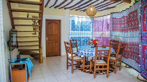 A dining area with a wooden table and chairs, a laptop on a small table, and a staircase with a wooden railing. - Charming 1-bed Cottage in Diani Beach 10min to bea (Diani Beach)