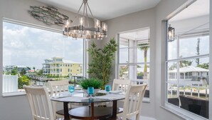 A dining area with a round table, white chairs, and a chandelier. There is a large window with a view of buildings and greenery outside. - 122 Primo Dr 3 Bedroom Home by RedAwning (Fort Myers Beach)