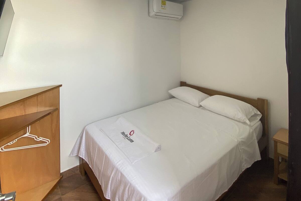 Un dormitorio pequeño y ordenado con una cama individual, un tocador de madera, un aire acondicionado de pared y una mesita de noche.