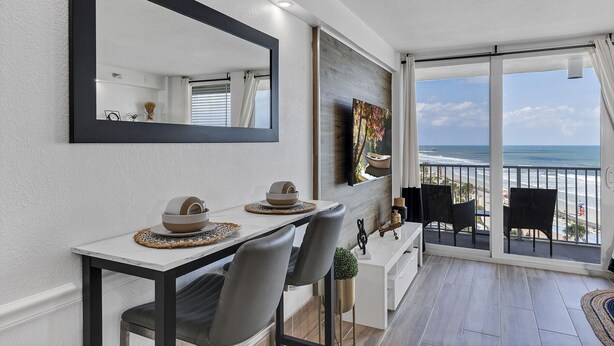 A modern dining area with a large mirror, a white table, two chairs, and a view of the ocean through a glass door. - Beachfront Remodeled Studio in Daytona Beach with King-Size Bed (Daytona Beach)