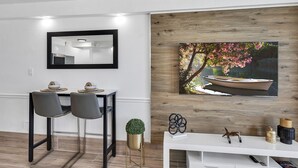 A modern dining area with a white table, two grey chairs, a black-framed mirror, and a wooden wall with a framed painting of a boat and flowers. - Beachfront Remodeled Studio in Daytona Beach with King-Size Bed (Daytona Beach)