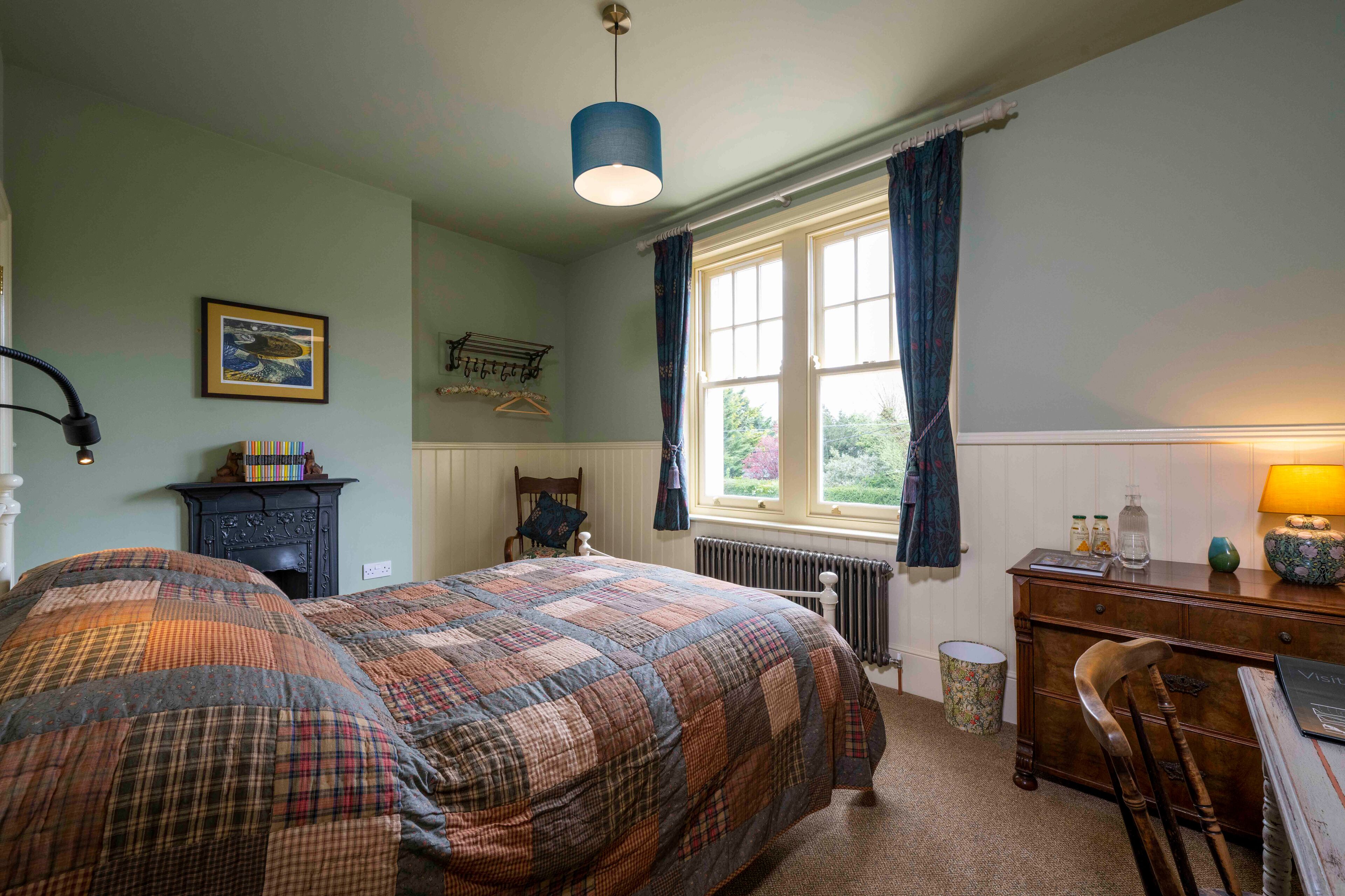 Une chambre à coucher avec un grand lit, une commode, une chaise, un radiateur, une fenêtre avec des rideaux et un luminaire suspendu.