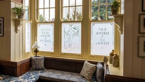 A room with a large bay window, a bench with patterned cushions, and framed artwork on the windowsills. - Long Man Inn (Polegate)