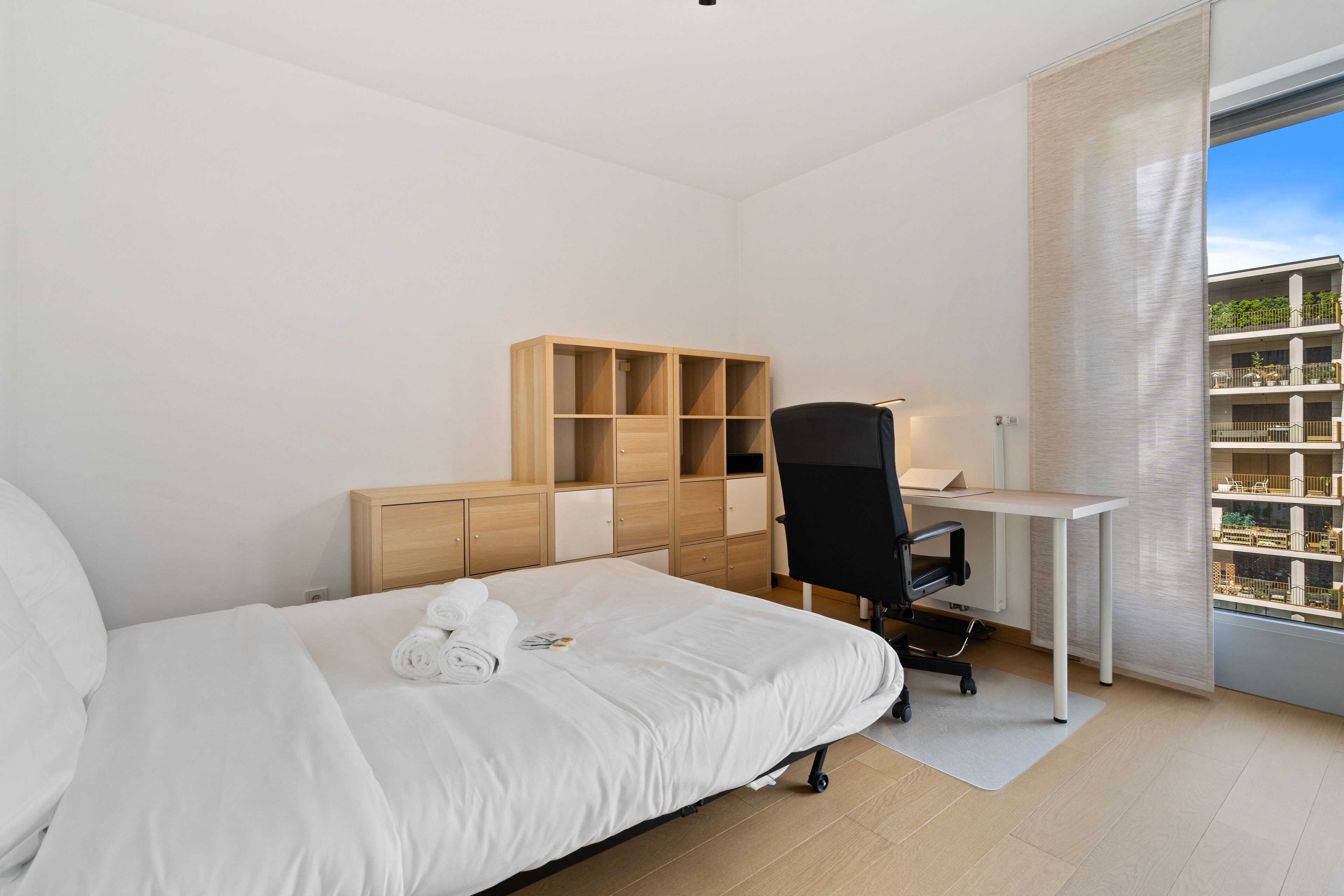 Une chambre d’hôtel moderne avec un lit bien fait, un bureau avec une chaise et une grande fenêtre donnant sur des immeubles.