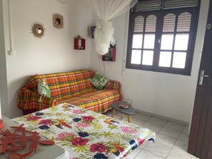 Une pièce comprenant un canapé à carreaux, une petite table, un lit recouvert d’une courtepointe à motifs floraux, une fenêtre ornée de volets décoratifs et des éléments de décoration murale.