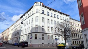 Monikerroksinen rakennus, jossa on kulma-sisäänkäynti, parkissa olevia autoja ja kirkas taivas.