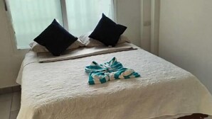 Un lit bien fait, recouvert d’une couvre-lit blanche et agrémenté de deux oreillers noirs, une serviette pliée, et une fenêtre avec des stores en arrière-plan.