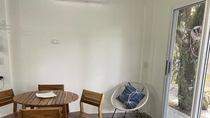 Un coin repas avec une table et des chaises en bois, une chaise blanche avec un coussin à motifs et une porte-fenêtre coulissante donnant sur un espace extérieur.