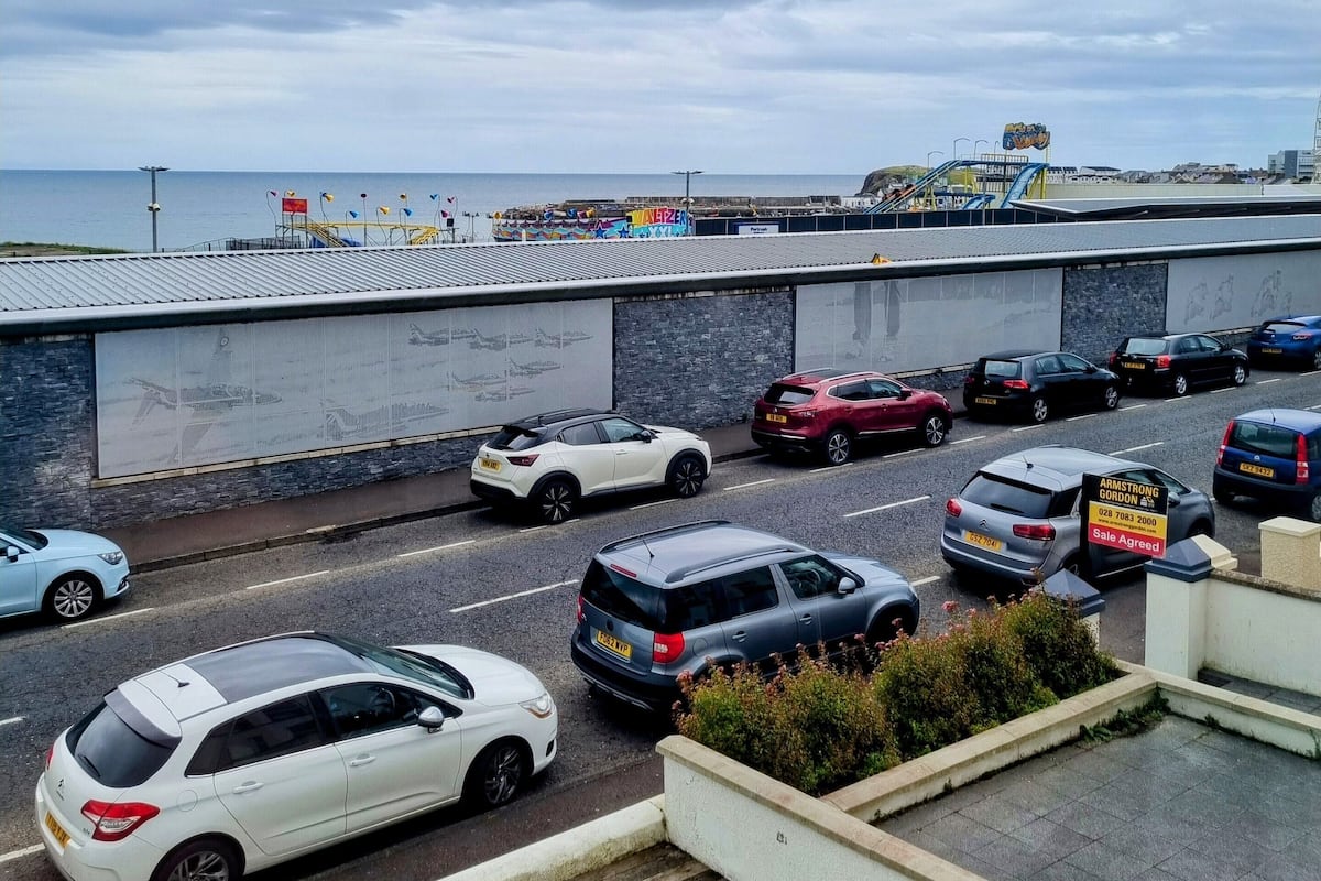 A parking area with multiple cars, a building with a stone facade, and a view of the sea in the background.